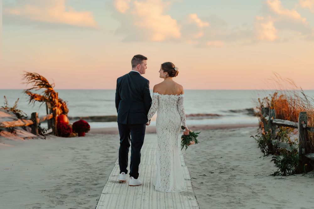 Bride and groom walking hand in hand on a beach boardwalk at sunset with floral arrangements nearby.