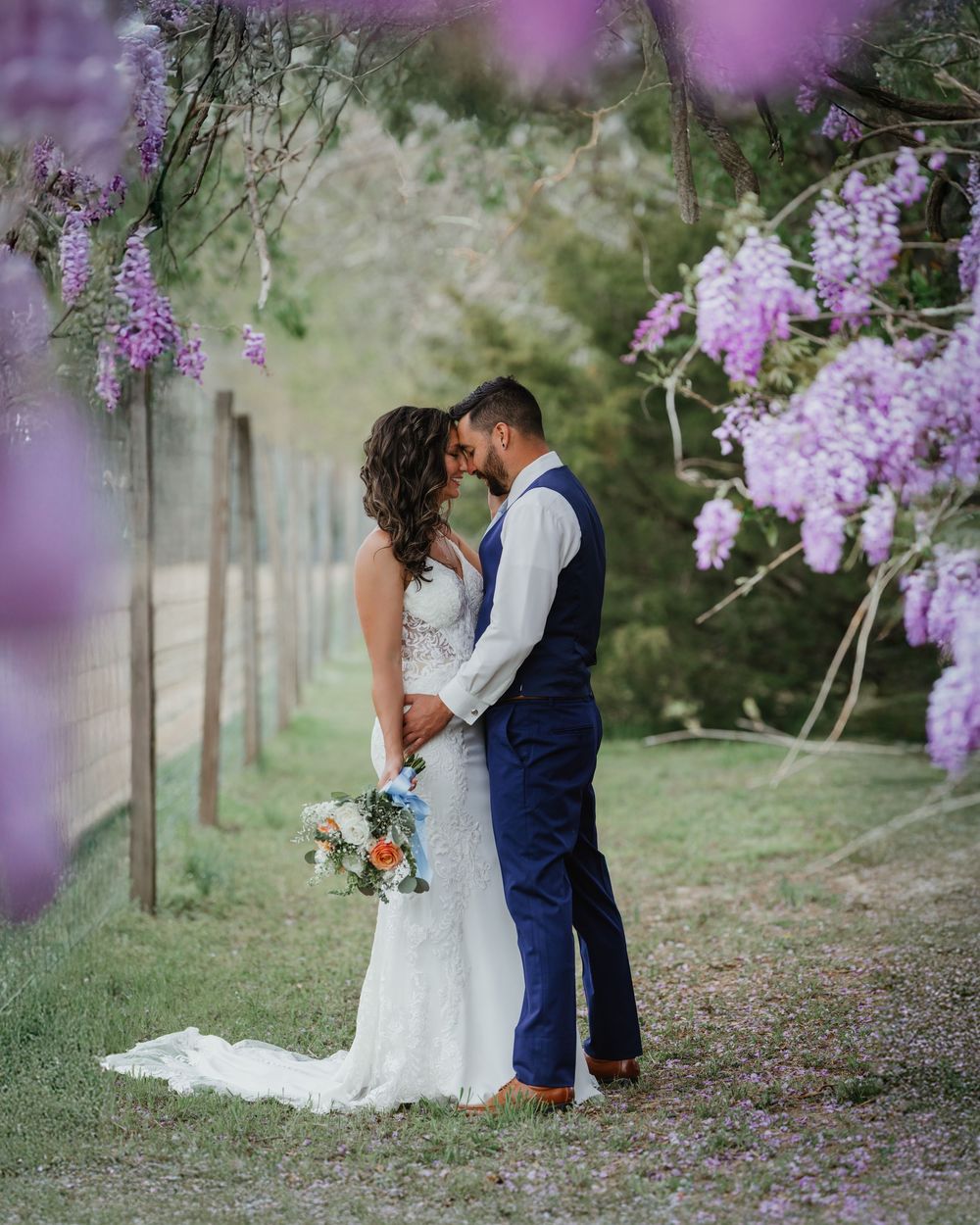 Bride and groom sharing a romantic kiss under blooming purple wisteria trees in a lush garden setting.