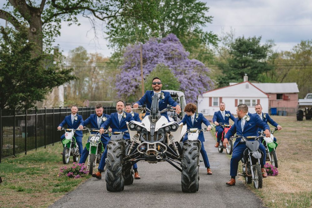 Groom and groomsmen in navy suits posing with ATVs and dirt bikes on a tree-lined driveway.