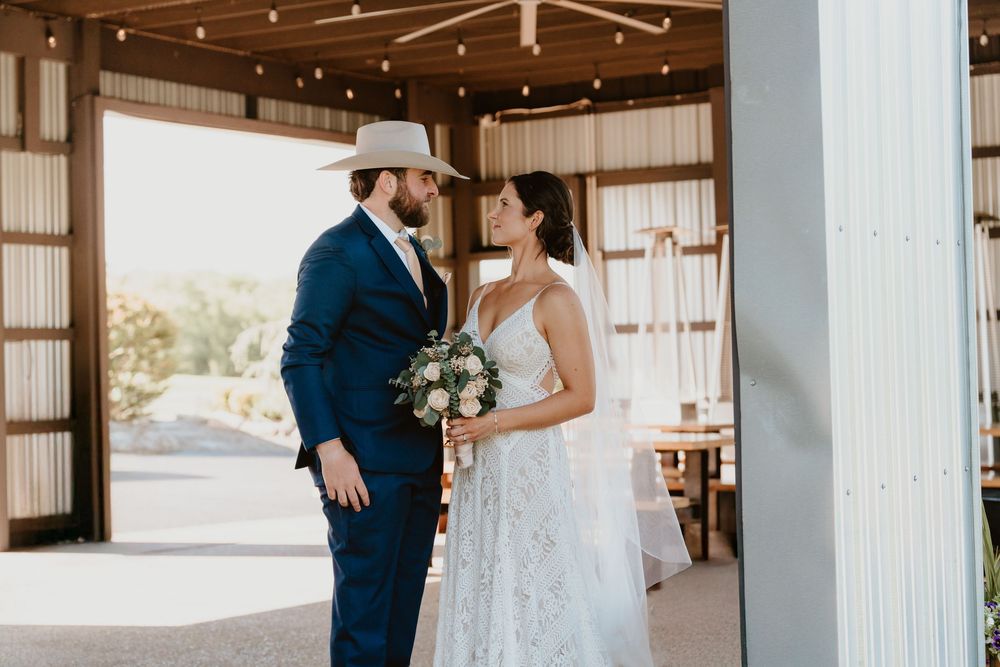 Bride and groom share a tender moment in a rustic barn wedding venue, groom wearing a navy suit and cowboy hat.