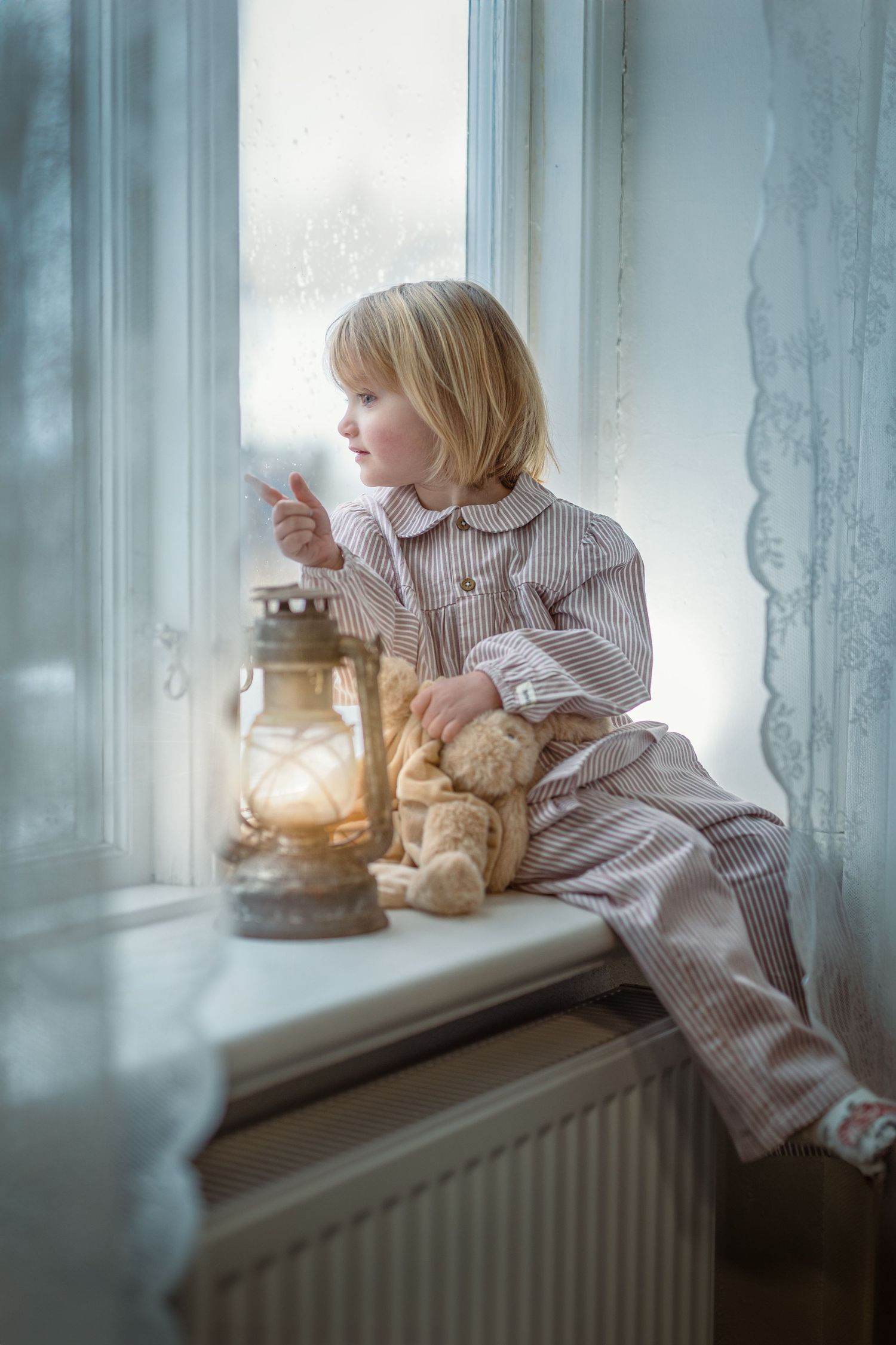 En liten flicka i randig pyjamas sitter i fönstersmygen. Hon tittar ut genom fönstret. Bredvid sig har hon sin nalle och en stormlykta.