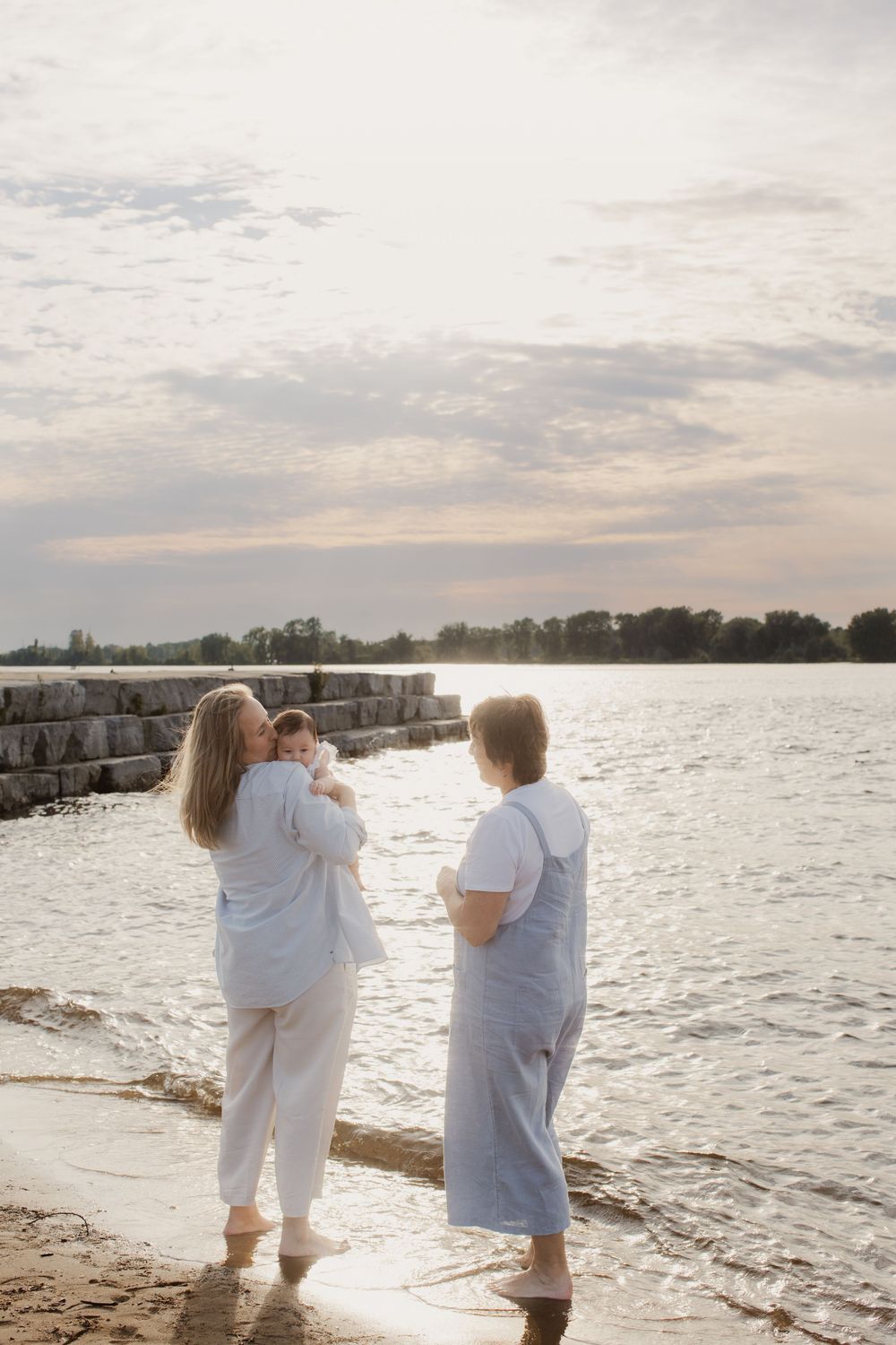 People wearing white outfits stand on a rocky beach at sunset overlooking a tranquil lake with cloudy skies.