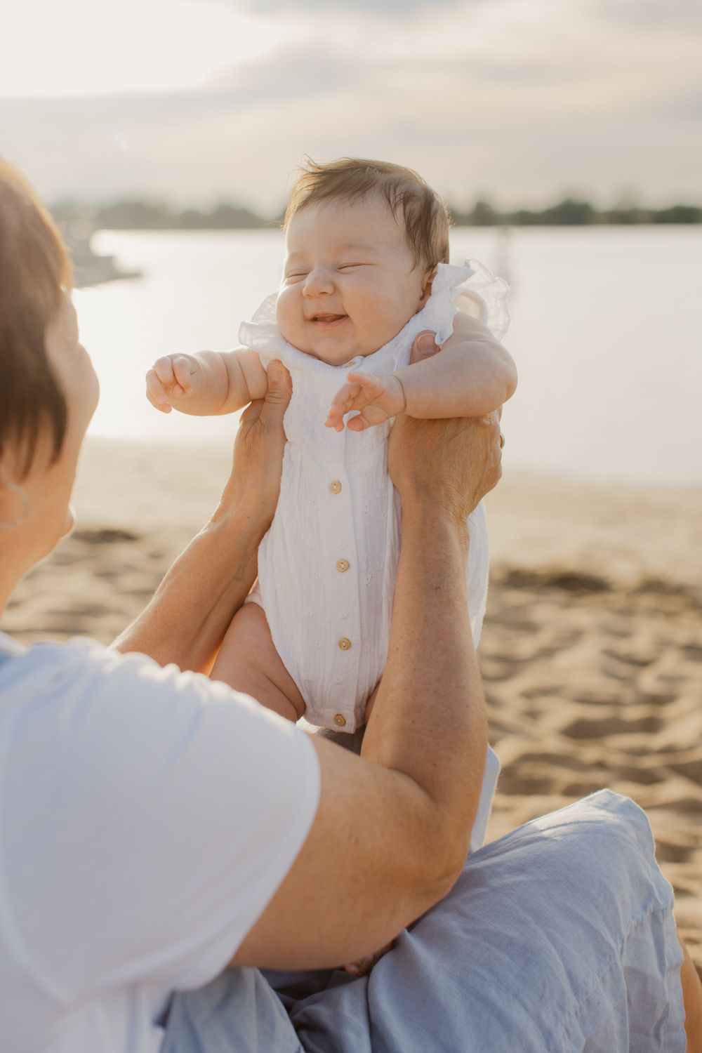 Happy baby in white onesie held up at sunset on sandy beach with parent in foreground.