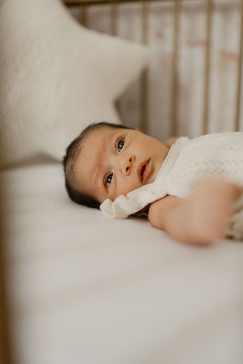 A tiny baby wearing a white outfit lays peacefully on soft white bedding while gazing at the camera.