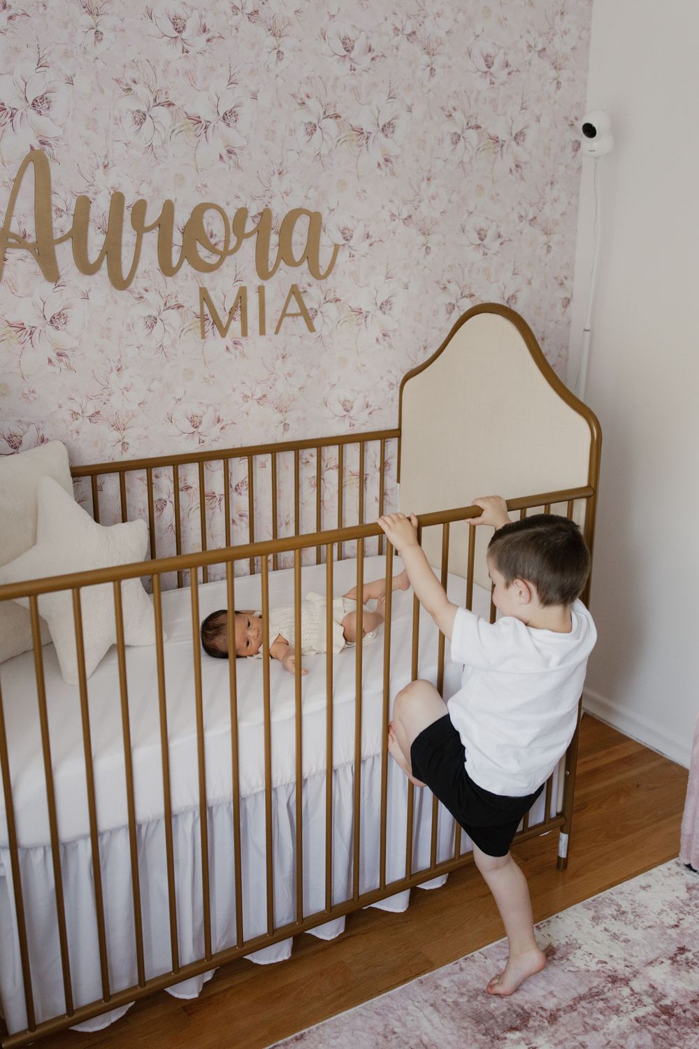 Gold text reading 'Aurora Mia' decorates a nursery wall above a white and gold crib with vintage floral wallpaper.