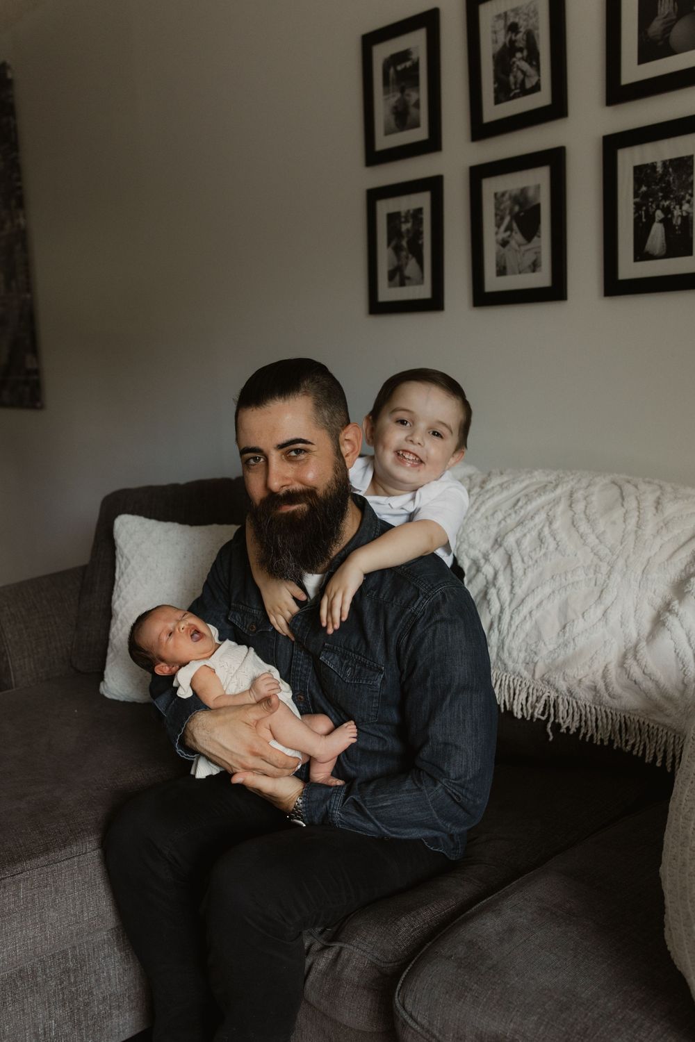 A loving moment captured on a couch with a newborn and toddler in a cozy living room with framed photos on the wall.