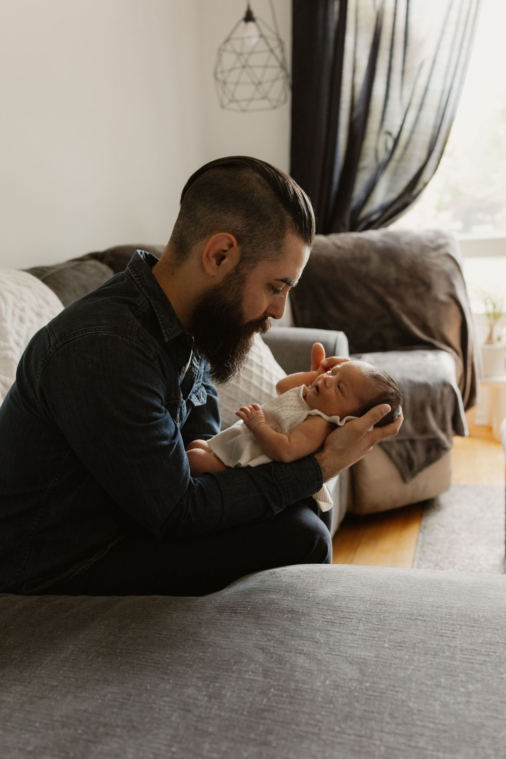 A bearded person sitting on a gray couch tenderly holds and gazes at a tiny baby in a warm, naturally lit living room.