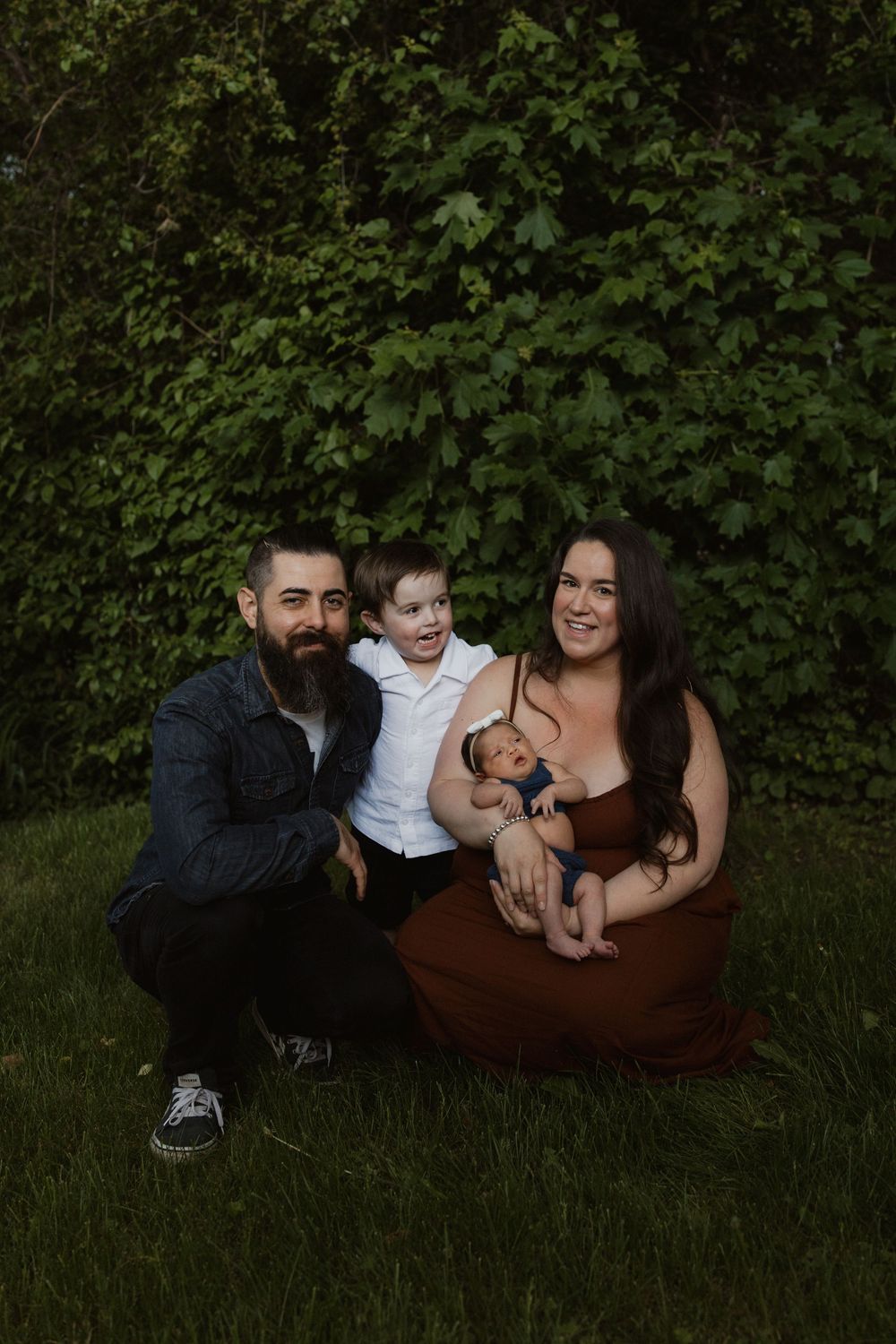 A family sits together on grass against a leafy green backdrop during an outdoor photo session.