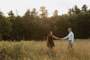 A romantic couple holds hands while walking through a golden meadow at sunset surrounded by pine trees.