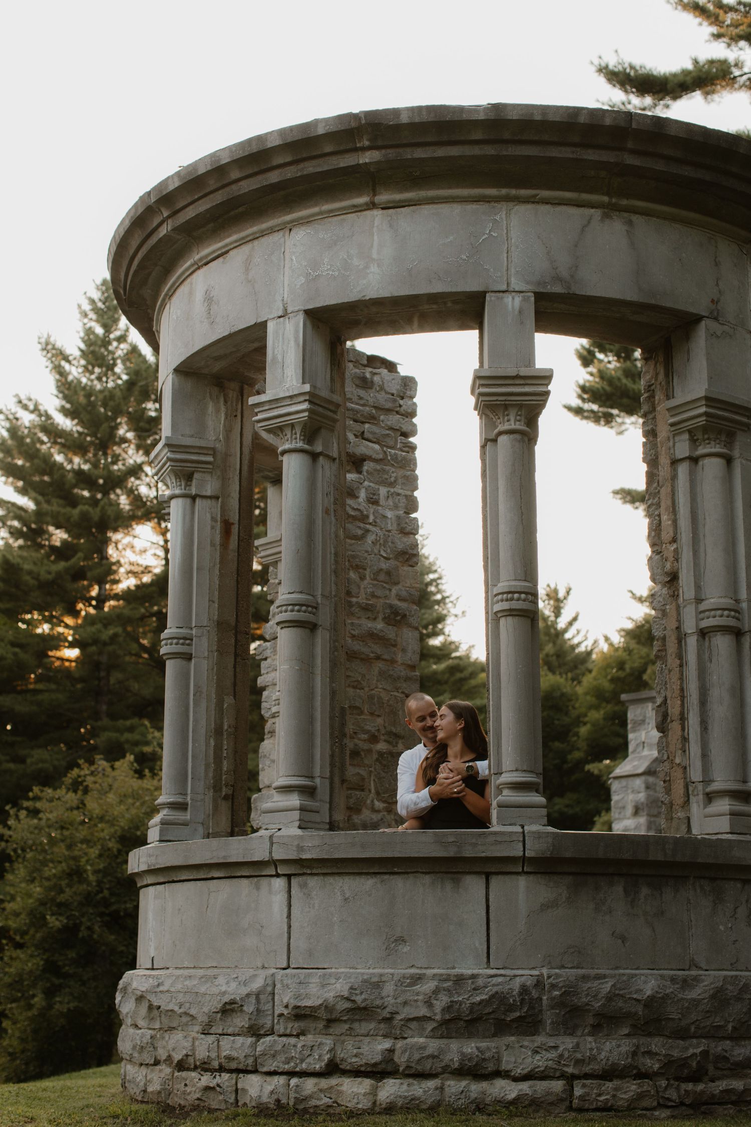 A couple embraces within a circular stone gazebo structure surrounded by evergreen trees at sunset.
