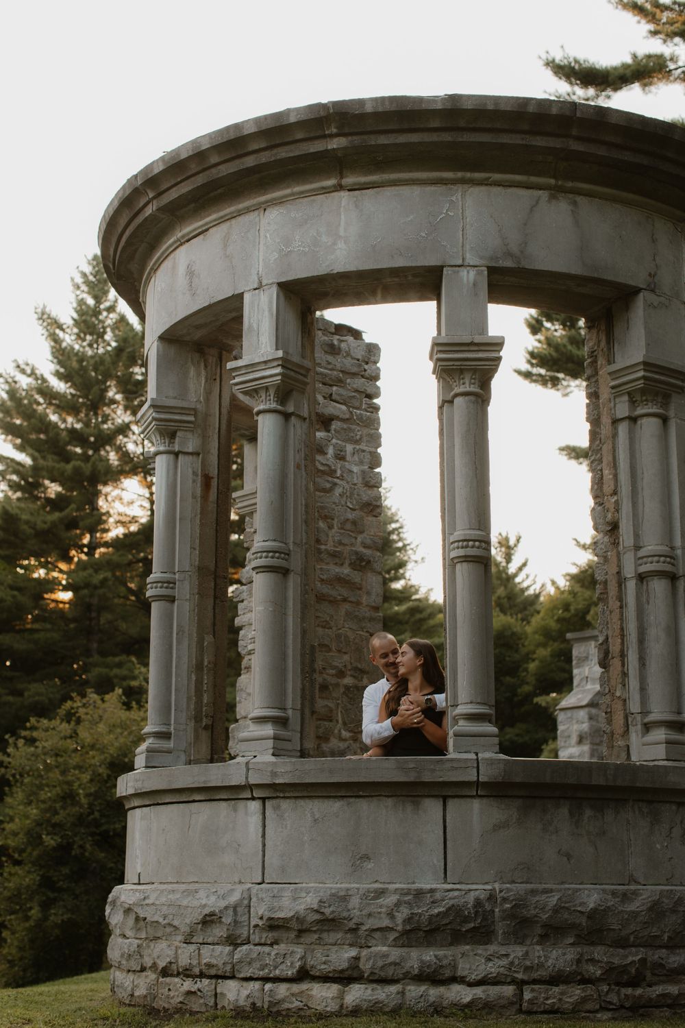 A couple embraces within a circular stone gazebo structure surrounded by evergreen trees at sunset.