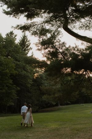 A couple walks hand in hand across a grassy lawn at sunset with towering pine trees silhouetted in the background.