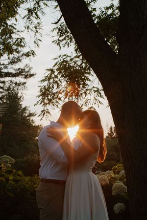 A couple embraces at sunset under a majestic tree with sunlight streaming through the branches creating a romantic silhouette.