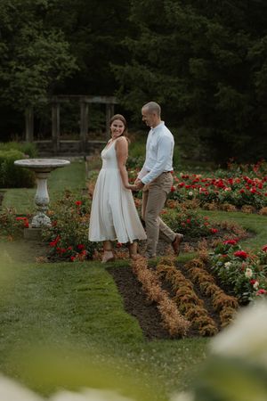 A couple walks together in a garden path surrounded by red roses and a stone birdbath on a summer evening.