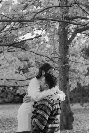 A romantic couple embracing outdoors in a black and white photo with bare tree branches above them.