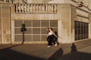 Silhouettes of people running past a building with shuttered windows in golden sunlight.