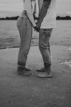 Two pairs of bare feet standing close together on a beach, wearing rolled up jeans in black and white.