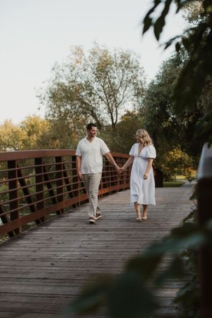 A couple holds hands while walking along a rustic wooden bridge surrounded by trees at sunset.