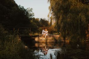 A couple sits together on a bridge over calm water at sunset with willow trees framing the scene.