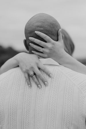 Black and white romantic photograph showing hands with rings embracing in a tender moment against a textured sweater.