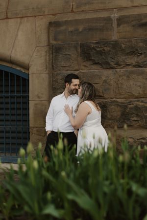 A romantic moment between a couple in elegant white attire standing against a stone wall with greenery in the foreground.