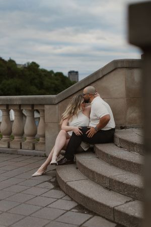 A romantic engagement photo session shows a couple reclining on stone steps with white columns at sunset.