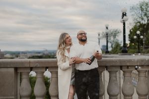 A romantic couple embraces on a scenic terrace overlooking a city, with ornate stone railings and lamp posts in the background.
