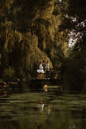A couple stands on a bridge over a serene pond with drooping willow trees.
