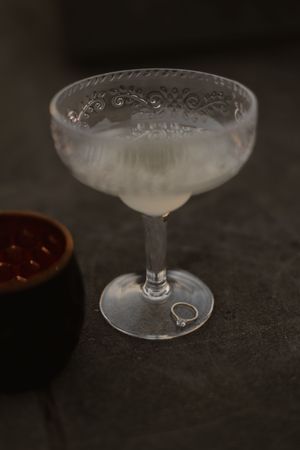 A frosted vintage coupe glass with elegant etched pattern sits next to a dark bowl on a moody surface.