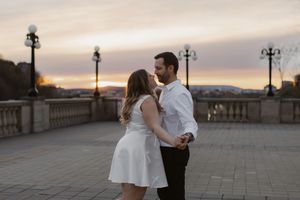 A romantic couple dancing on a stone bridge at sunset with ornate lampposts in the background.