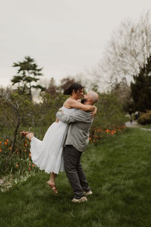 A couple embraces in a romantic dance pose on a grassy lawn with spring flowers and trees in the background.