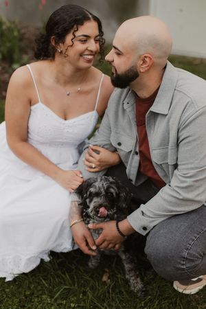 A couple in wedding attire share intimate moments with their small black dog on grassy ground during an outdoor photoshoot.