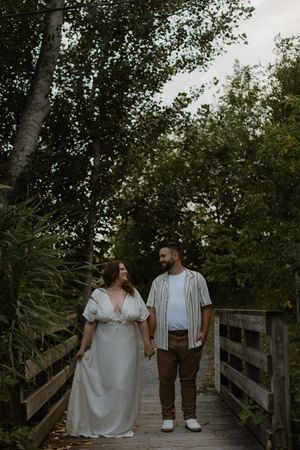 A couple walks together on a wooden dock pathway surrounded by lush green trees in a romantic woodland setting.
