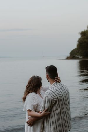 A couple embraces while looking out at a calm lake with tree-lined shores under an overcast sky.