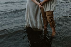 A couple in light summer clothing stands together at the edge of dark rippling water during sunset.