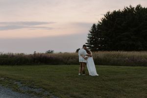 Couple in elegant attire walks across a scenic hilltop meadow at sunset for wedding portraits.