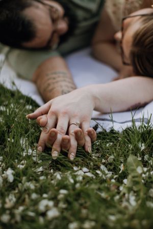 Close up of intertwined hands with rings lying in grass with white flowers.