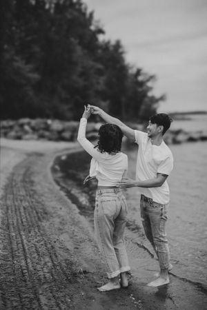 Black and white photo of couple dancing on beach near rocky shoreline.