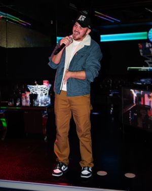Performer in denim jacket and khakis singing on stage at nightclub venue.