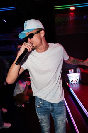 Performer in baseball cap and white t-shirt singing into microphone at club venue.