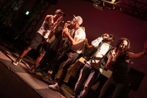 Performers on stage singing into microphones against a red backdrop during a live music performance.