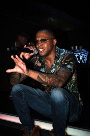 An energetic performer in sunglasses and a tropical print shirt crouches while singing into a microphone on stage.