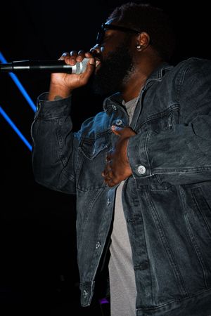 A performer in a dark denim jacket sings passionately on stage with blue lighting in the background.