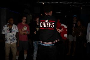 Person wearing a Chiefs jacket standing among others in a dimly lit room.