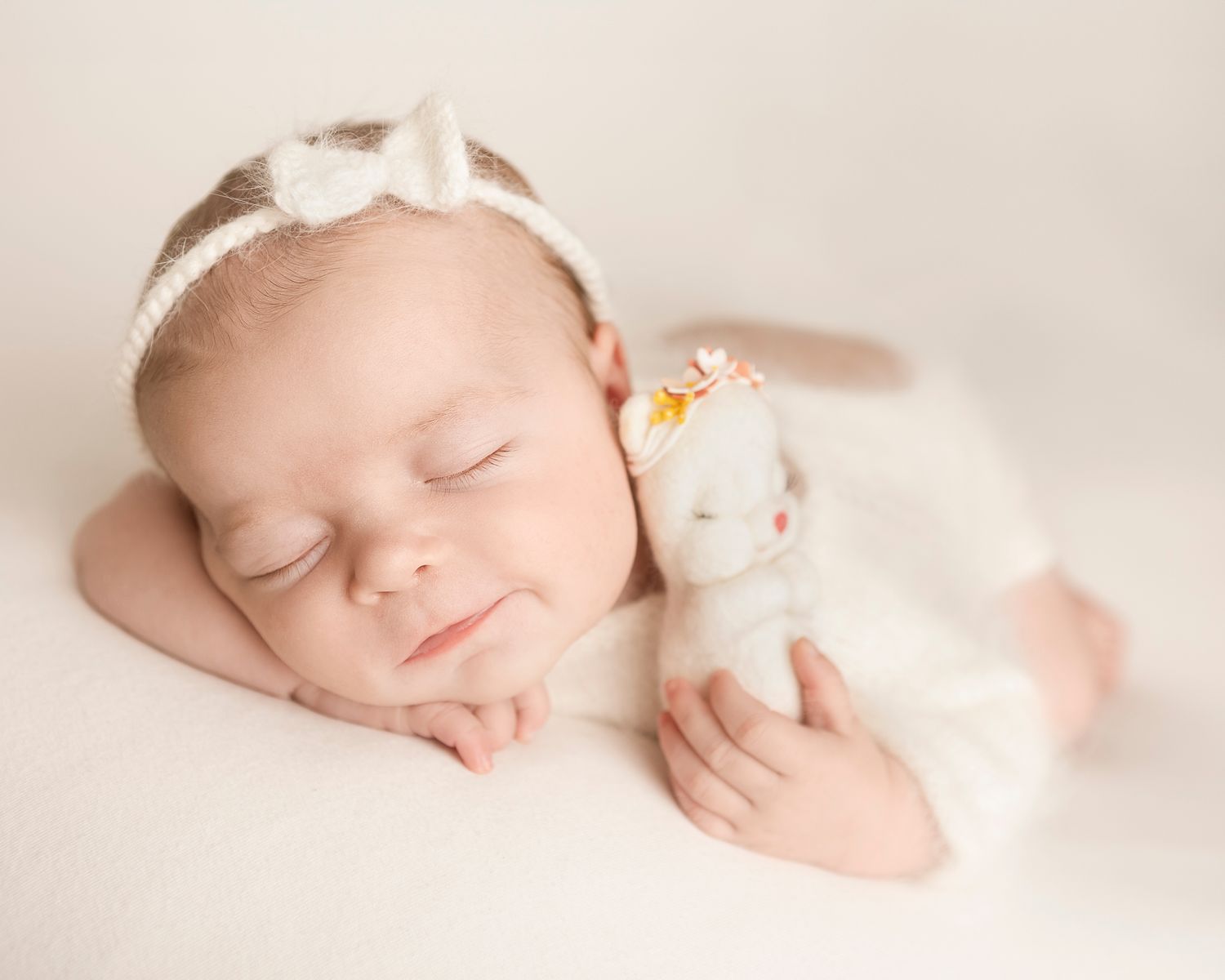 Baby på hvit bakgrunn med hvit body og hvit sløyfe på hodet. Lukkede øyne, smiler og holder en hvit bamse med blomsterbånd på hodet