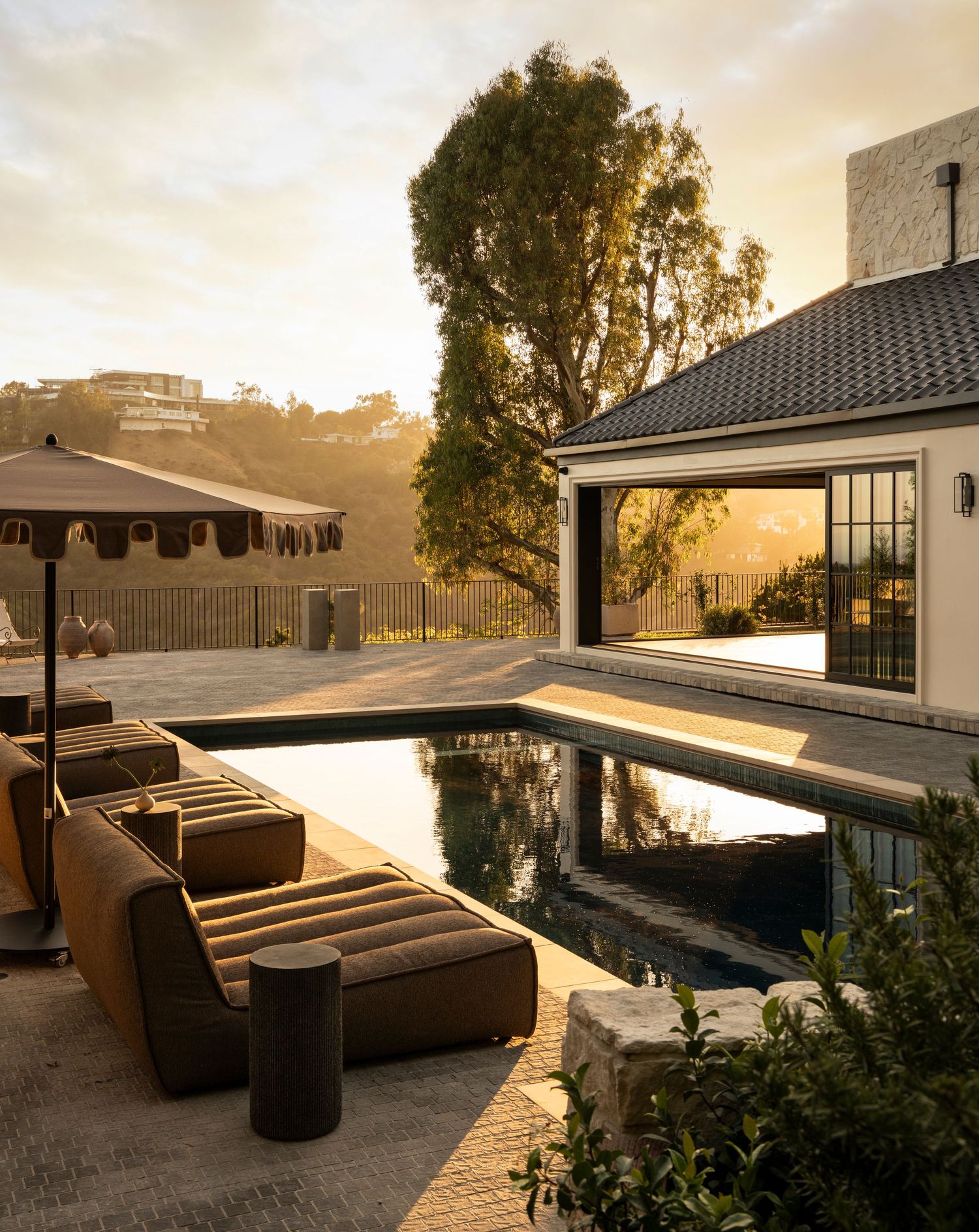 Luxury outdoor pool area with modern seating, patio umbrella, and glass-walled structure at golden hour sunset.