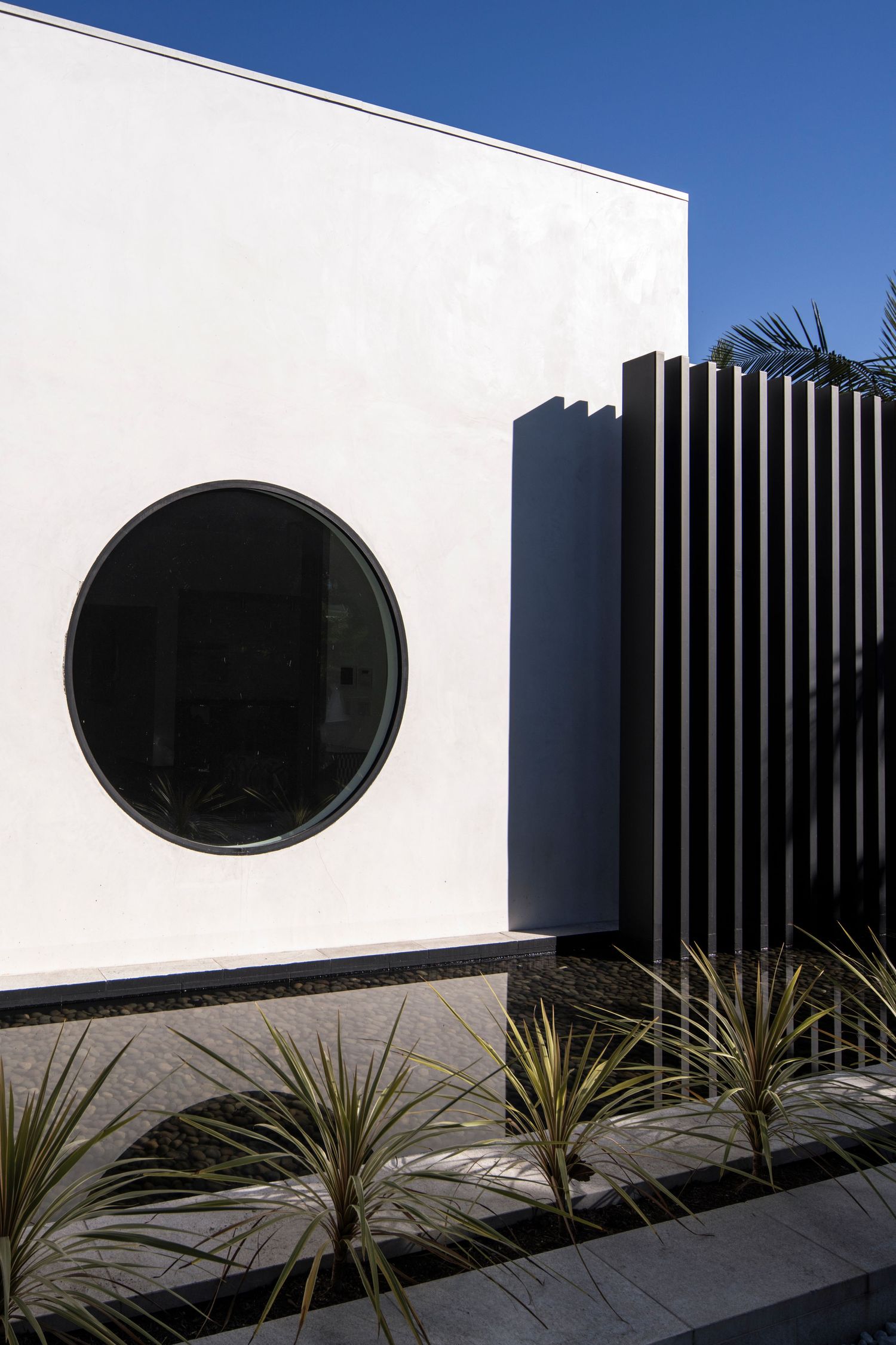 Modern architectural design featuring a white wall with circular window and vertical slat detail against blue sky.
