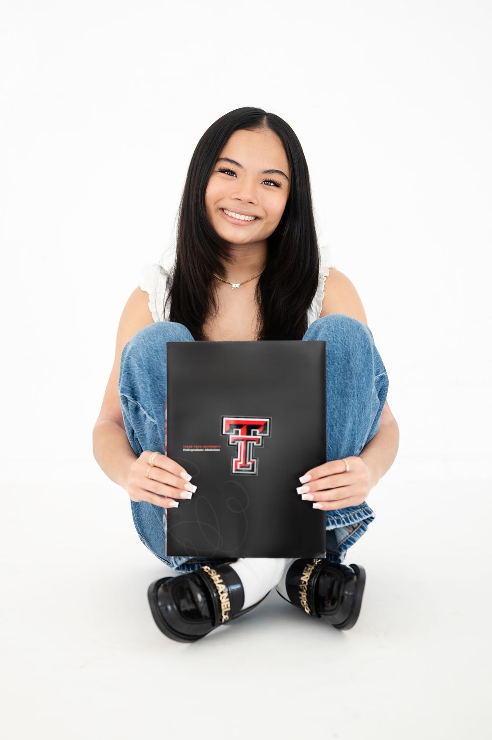 Person in denim holding Texas Tech logo portfolio while sitting cross-legged on white background.