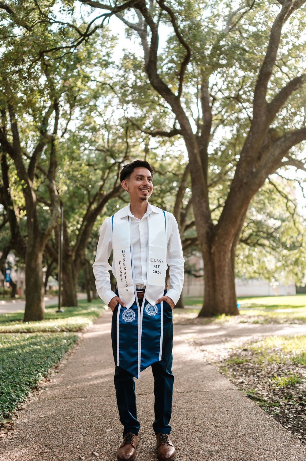 A smiling graduate wearing a white stole and Class of 2023 sash stands on a tree-lined campus pathway.