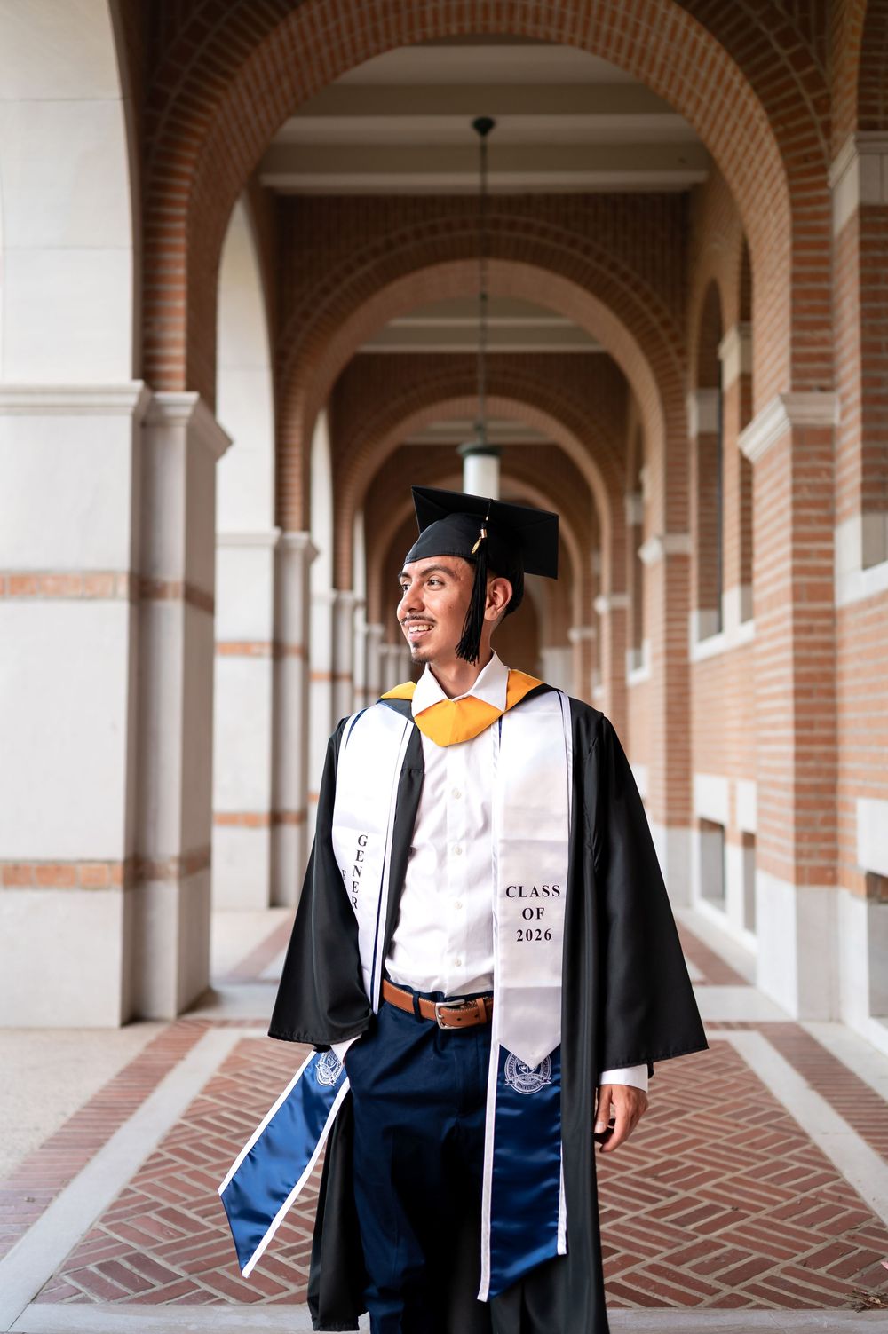 Smiling graduate in cap and gown with Class of 2020 stole walks through brick archway.